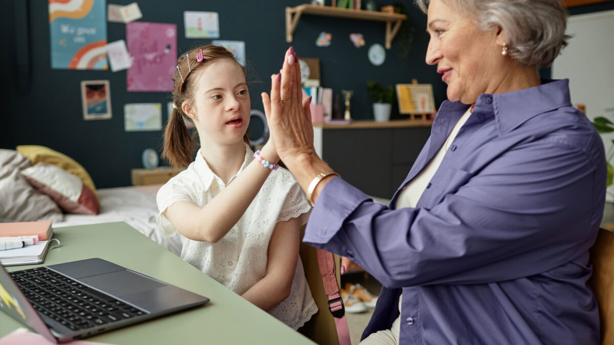 Elderly woman engaging with young girl with Down syndrome in cozy classroom setting high-fiving while spending time together with laptop