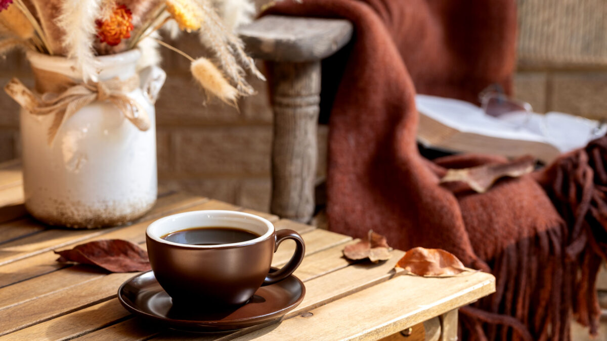 Cup of coffee with vase of dried autumn flowers and leaves on wooden table and old chair with blanket in background