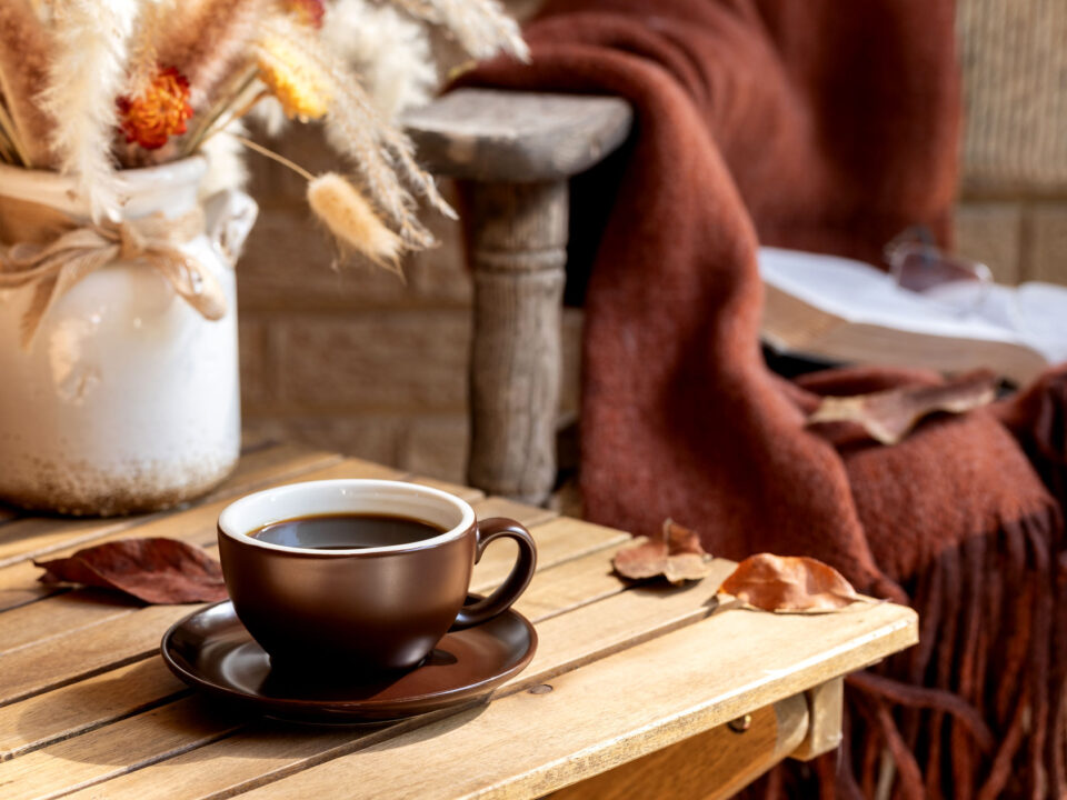 Cup of coffee with vase of dried autumn flowers and leaves on wooden table and old chair with blanket in background