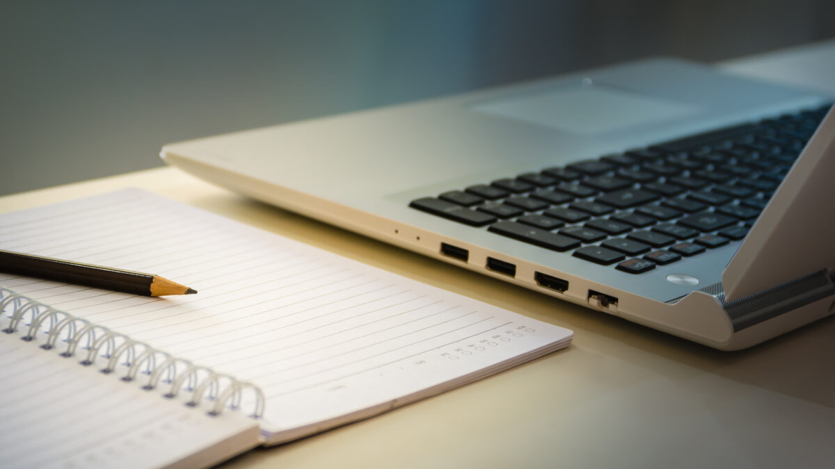 Office workplace with laptop and blank note book paper with pencil under warm light of desk lamp.