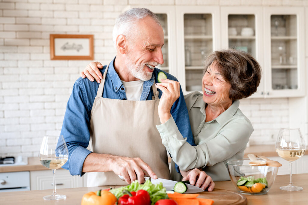 Cheerful old senior wife treating her husband while helping him cook prepare dinner salad lunch at home kitchen.