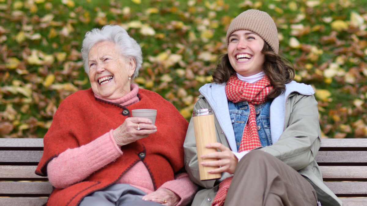 Portrait of granddaughter and grandmother sitting on bench in autumn park, drinking coffee, laughing together