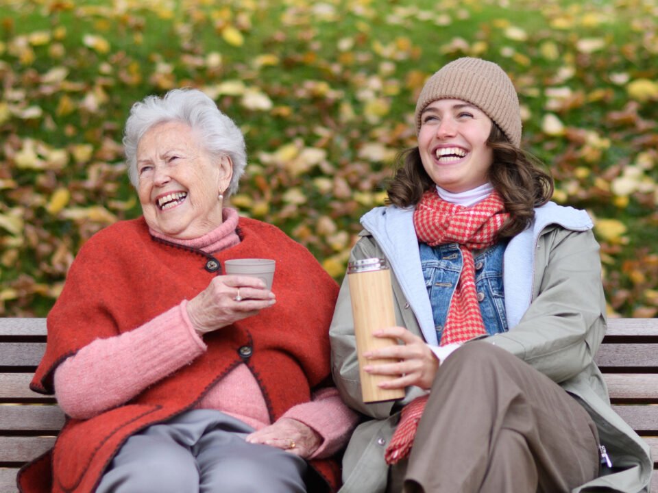 Portrait of granddaughter and grandmother sitting on bench in autumn park, drinking coffee, laughing together